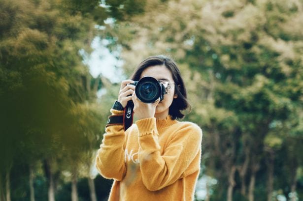 woman with yellow jacket holding camera with trees 