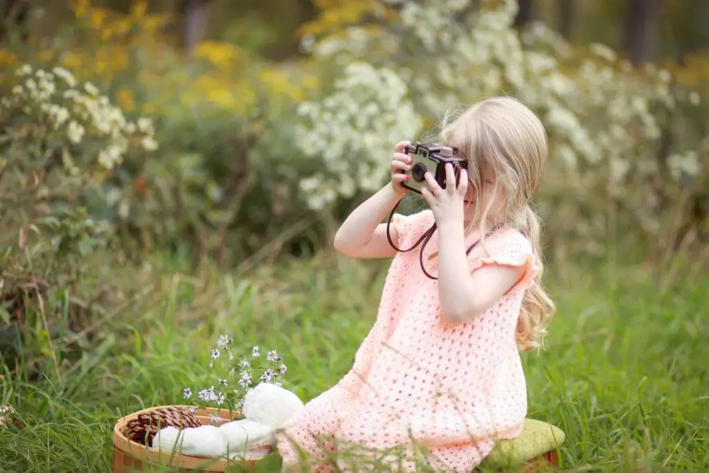 A little girl sitting on the grass, taking a picture with her camera