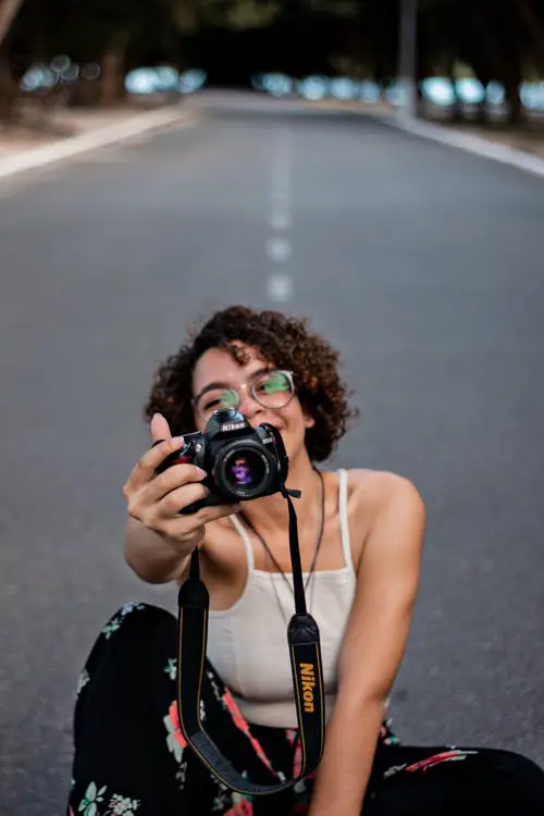 Woman wearing glasses and sitting on the center of the road while holding a camera