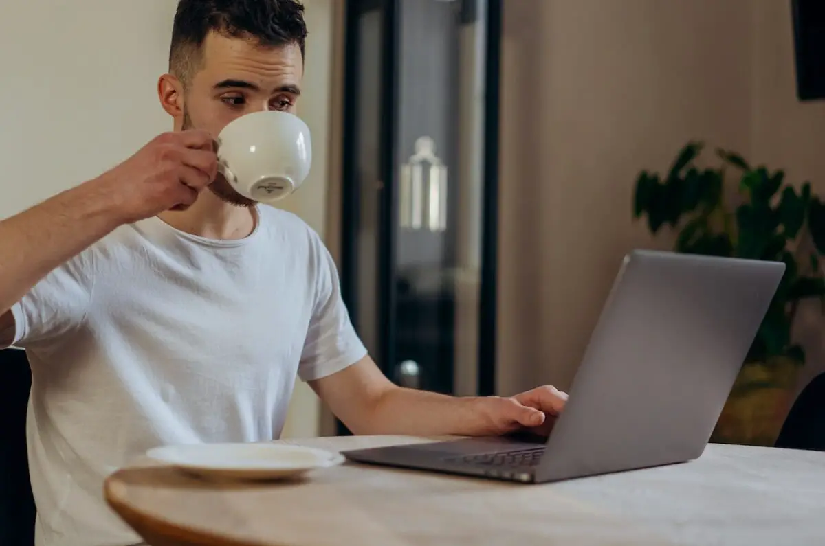 A man using a silver laptop and a white plate on a wooden table while drinking coffee in a white ceramic mug