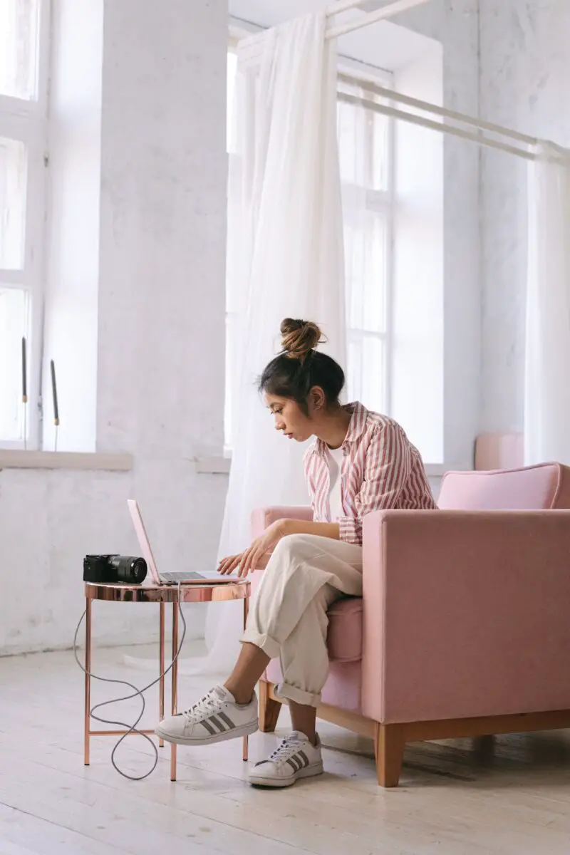 A DSLR camera connected to a pink laptop being used by a woman wearing a pink striped shirt sitting in a pink armchair