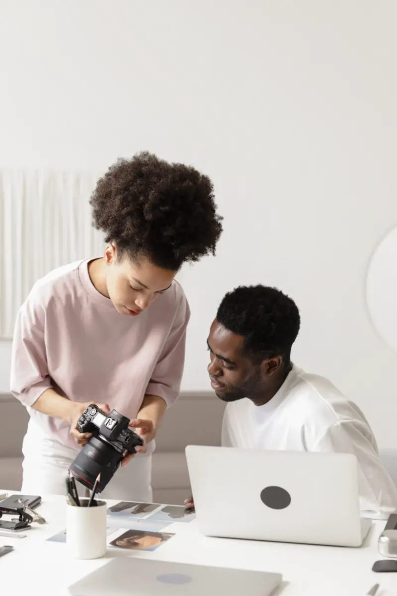 A young woman in a pink shirt showing a photo taken on a Canon DSLR camera to a man who is seated at a white table in a living room