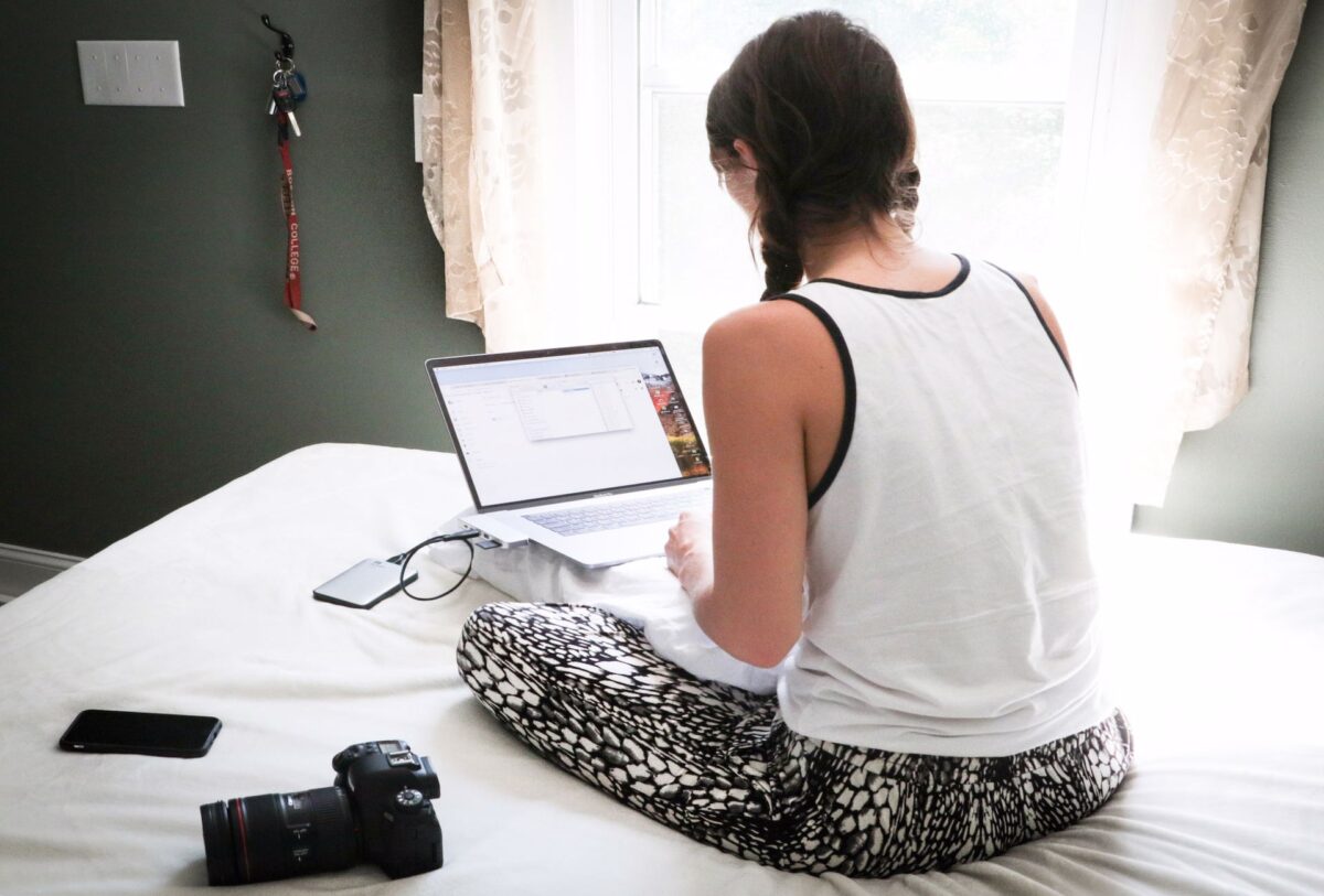 A woman using a silver laptop with a black external hard drive connected to it and a DSLR camera and smartphone on top of a white bed in the room